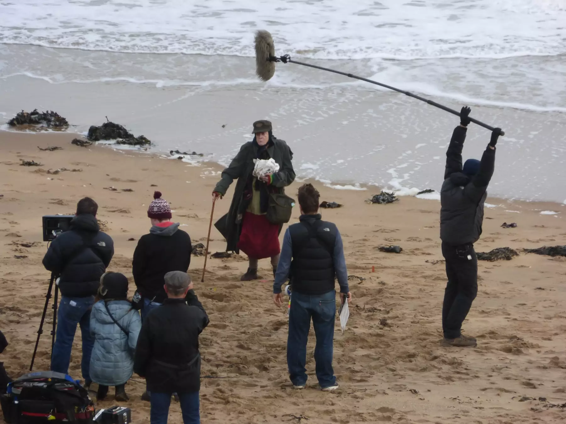 Actress stood close to waters edge holding papers and a walking stick. In front in an arch film crew of 6 including one with a pole microphone